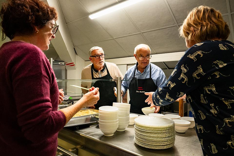 Deux hommes âgés avec des tabliers et deux femmes autours d'assiettes et d'un plat cuisine dans un réfectoire.