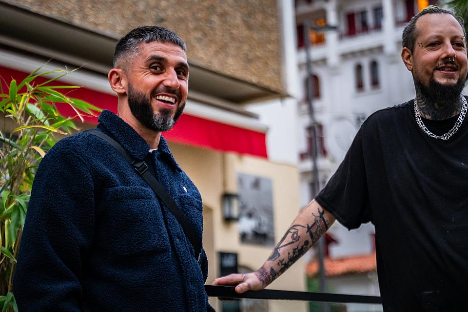 Un homme barbu souriant regarde l'objectif devant la devanture d'un restaurant. Un autre homme au bras tatoué se tient de bout près de lui et sourit aussi. 