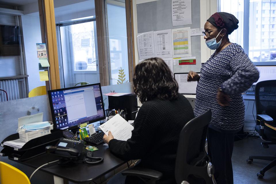 Deux femmes dans un bureau de profil devant un ordinateur. L'une est debout, l'autre assise, de dos, des papiers à la main. 