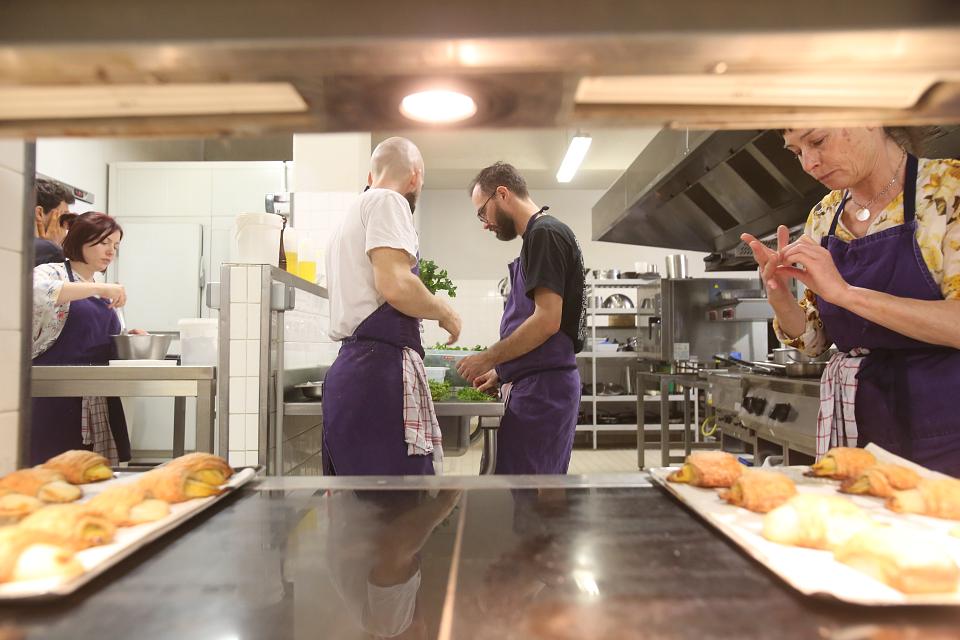 Cinq personnes échangent dans une cuisine collective, on voit des plats au premier plan.