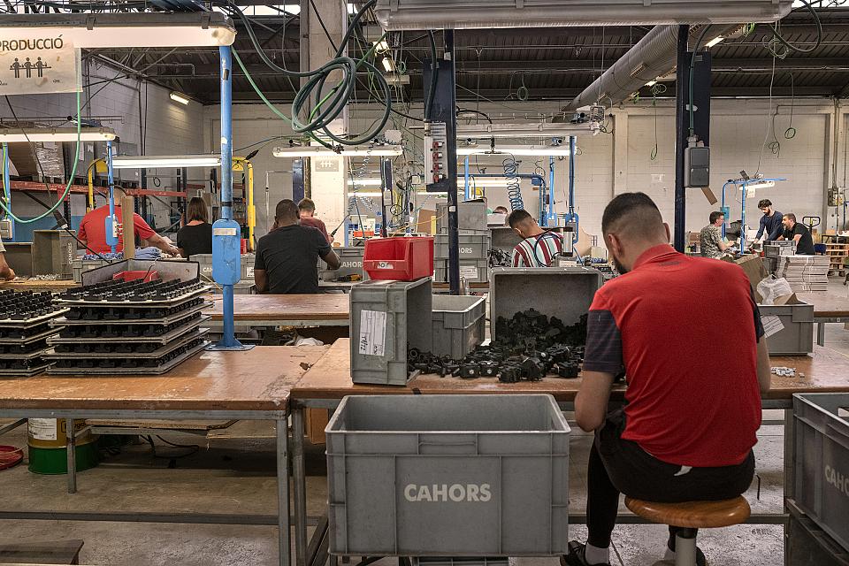Dans un atelier de production, des hommes de dos assis à des tables devant des pièces industrielles. Sur une caisse en plastique est écrit le mot « Cahors ».