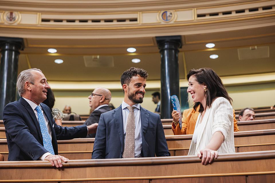 Un jeune homme en costume souriant entre une femme et un homme qui lui parlent, debout dans l'hémicycle d'une assemblée. 