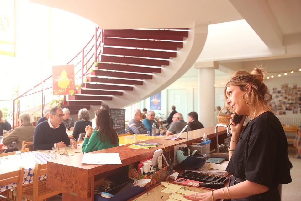 La salle du restaurant, des clients attablés au pied d'un large escalier en colimaçon, une femme à la caisse au téléphone au premier plan.