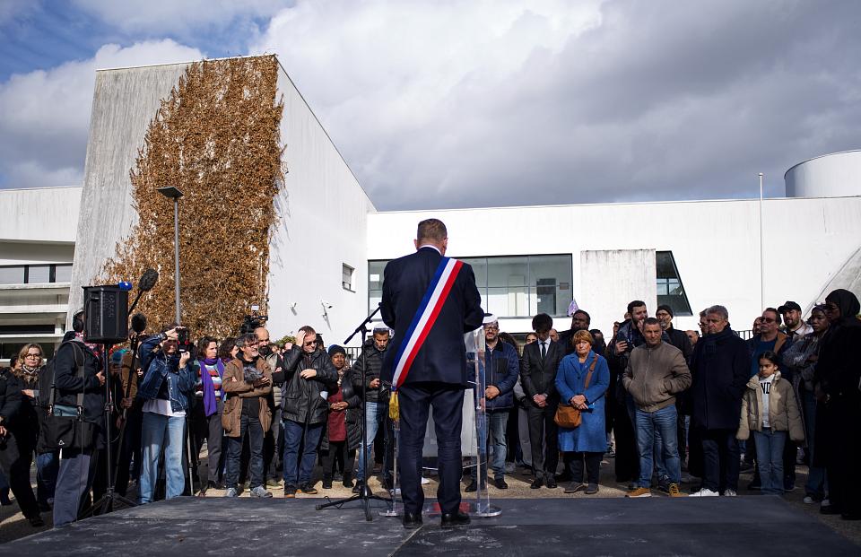 Un homme de dos qui porte une écharpe triclore parle devant une cinquantaine de personne debout. 