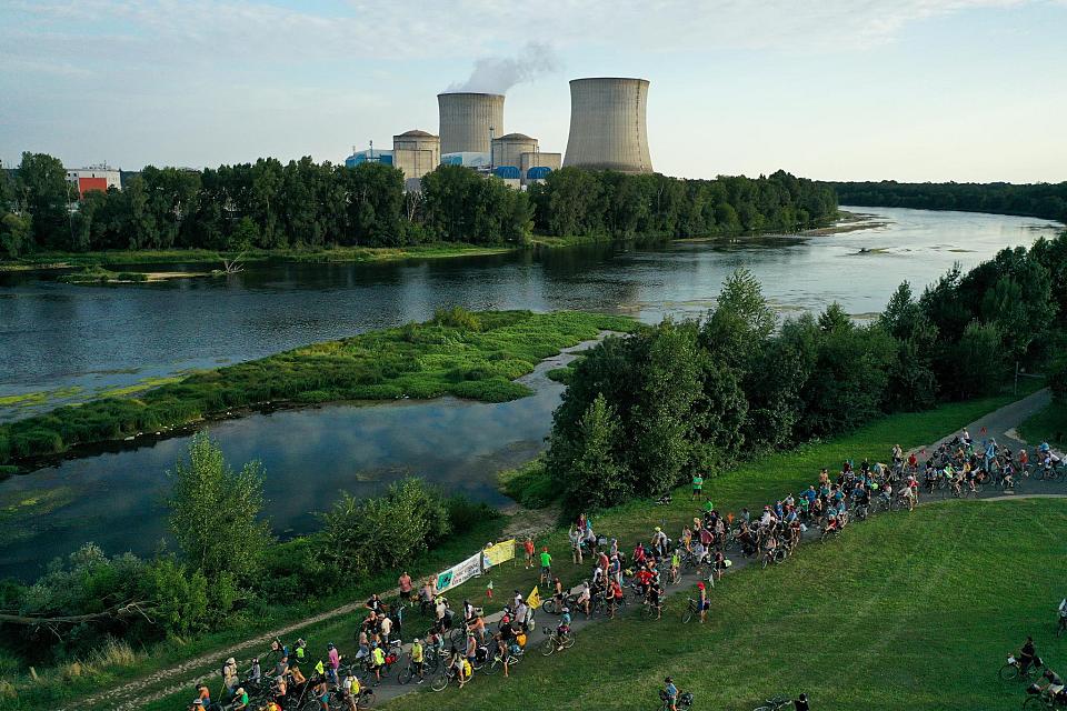 Au centre de l'image, un fleuve, avec à gauche une centrale nucléaire et à droite de très nombreux cyclistes arrêté·es devant deux banderoles.
