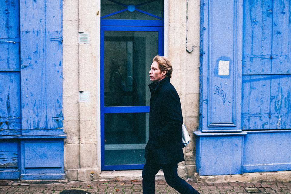 Wilfrid Séjeau de profil marche dans une reu edevant des murs bleus, il a les mains dans les proches. 