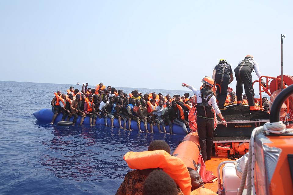 Des dizaines de personnes sur une embarcation de fortune avec des gilets de sauvetage et un autre bateau qui leur vient en aide. 