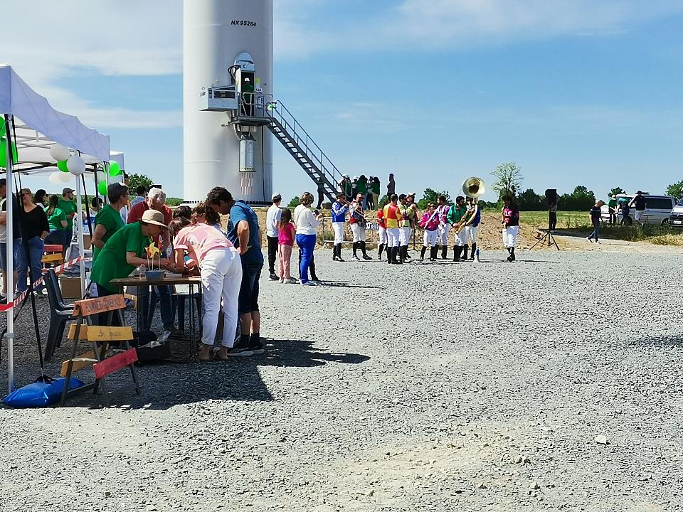 Une fanfafre au pied d'un mât d'éolienne au second plan, un stand avec des perosnnes autour au premier plan. 