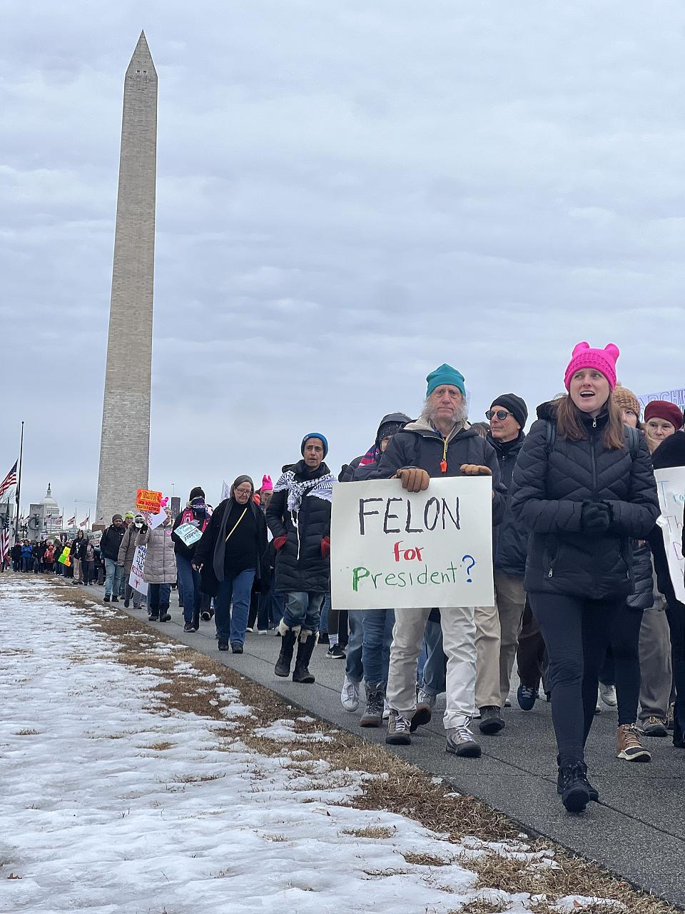 Des personnes défilent à Washington DC avec au fond l'obélisque du Washington Monument. Un homme tient une pancarte sur laquelle il est écrit « Felon for president ? »