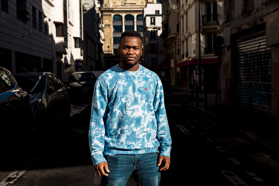Portrait d'Arnaud Coulibaly, debout dans la rue, en jean et pull bleu. 