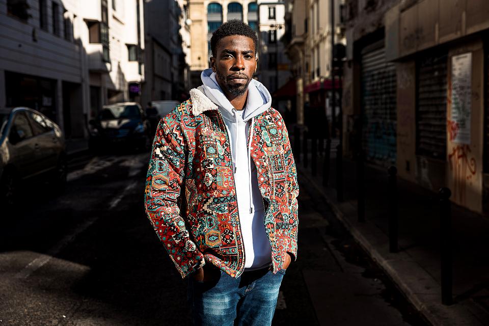 Portrait d'Ibrahima Cissokho, debout dans une rue, en jean, pull blancn et veste colorée. 