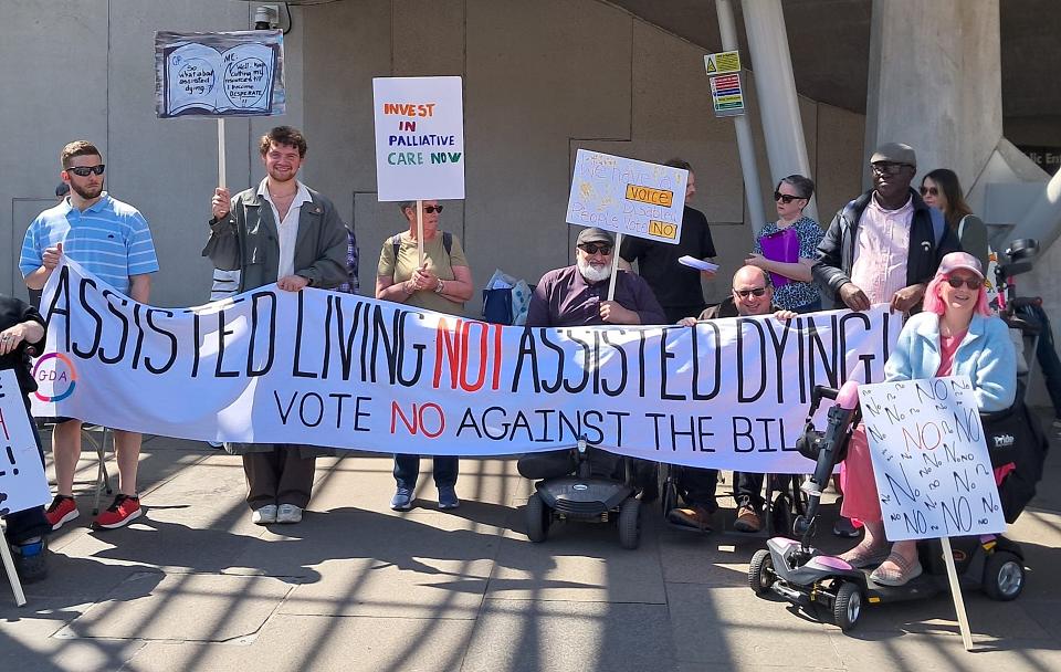 Des personnes, dont plusieurs en fauteuil roulant, portent une pancate "Assisted Living Not Assisted Dying".