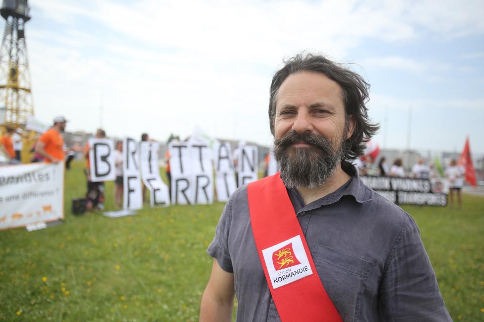 Un homme avec une écharpe rouge en travers de la poitrine sur laquelle ose trouve le blason de la Normandie regarde l'objectif. Derrière, dans un champs, un groupe de personne tiennent des lettres qui forment le mot « Brittany Ferries ».