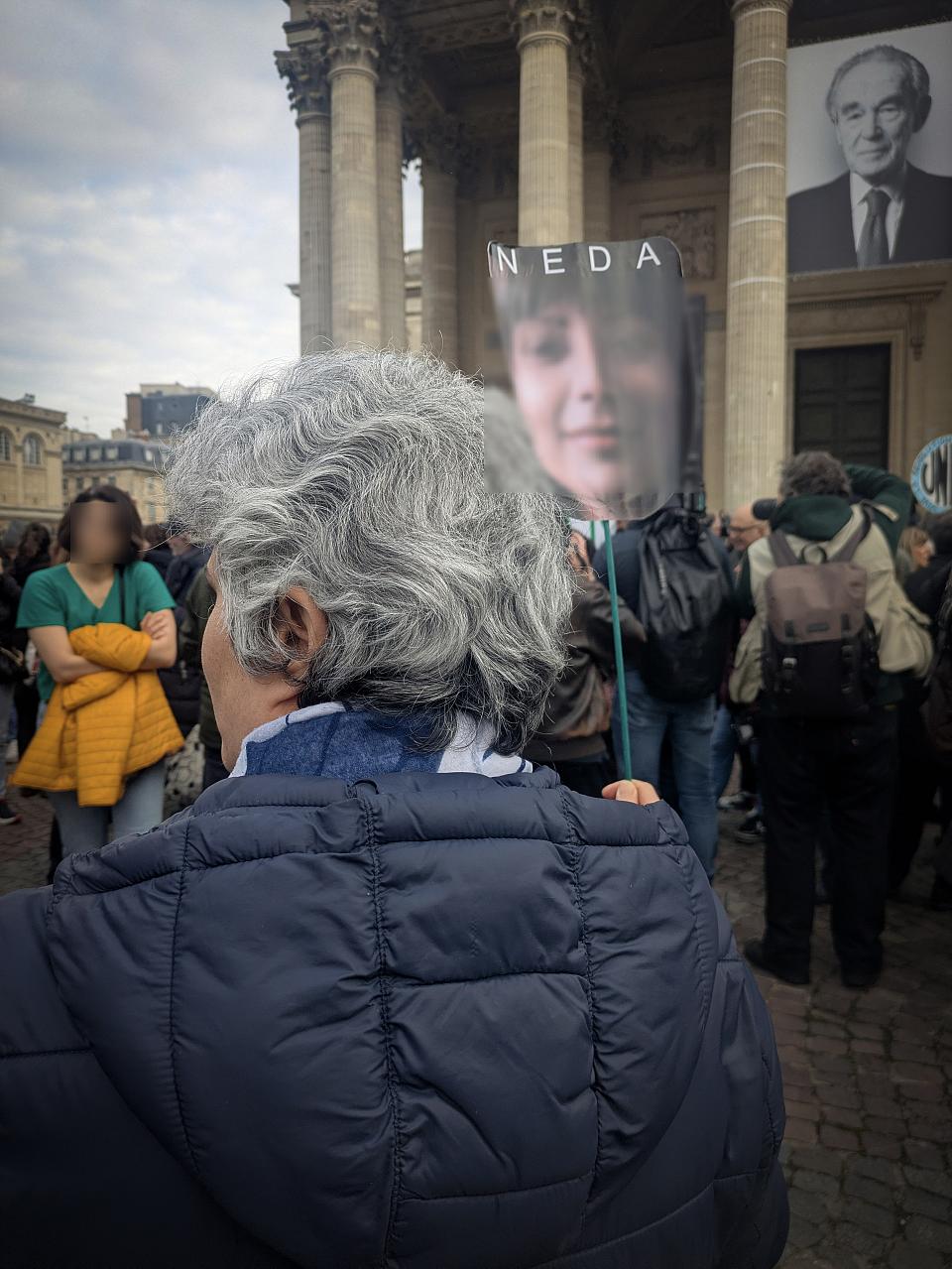 Une femme aux cheveux courts et gris vue de dos au milieu d'un rassemblement. 