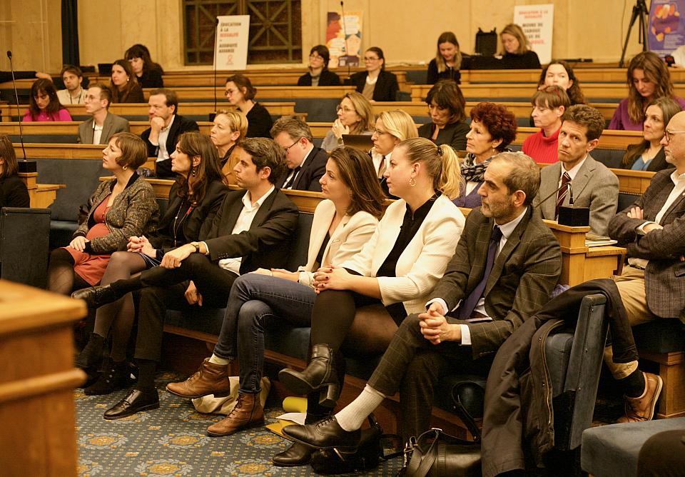 Une vingtaine de personnes assises sur les bancs en bois d'une petite salle. 