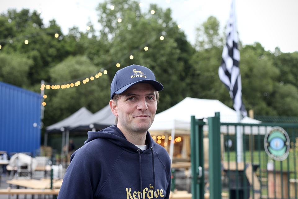 Erik Tegnér devant la brasserie Kerfave avant son inauguration, le 14 mai 2025. Il arbore une casquette et un sweat bleu marine au nom de la brasserie, qui est en arrière blanc, avec à droite un drapeau breton. 