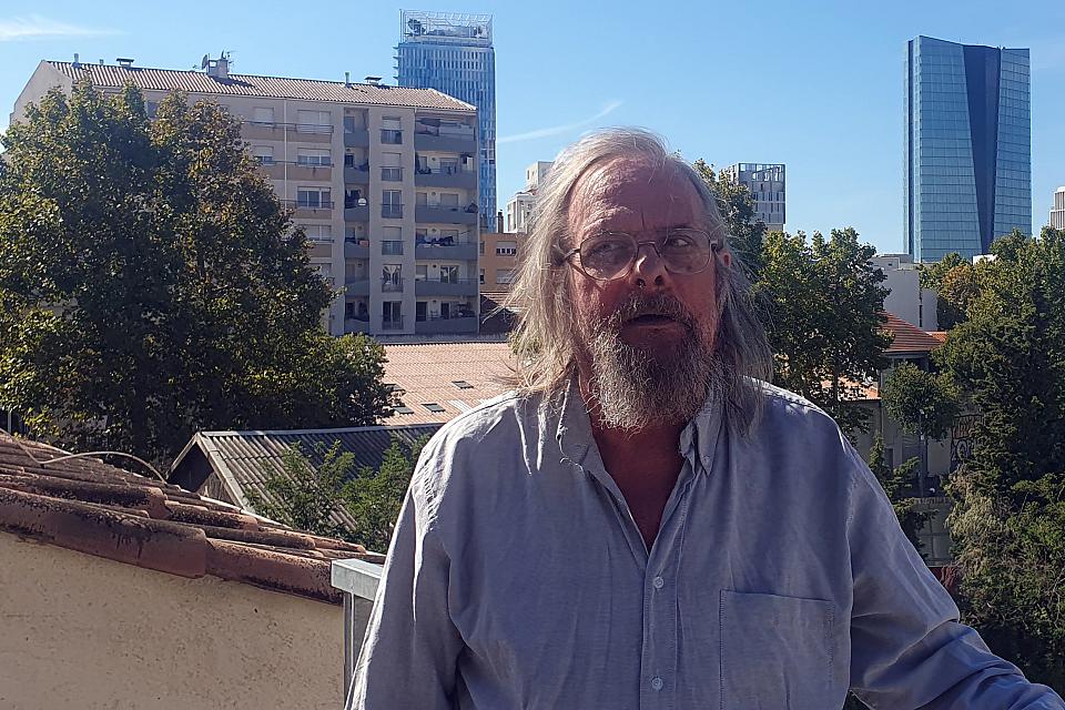 Un homme aux cheveux longs et blancs et barbu debout sur une terrasse.