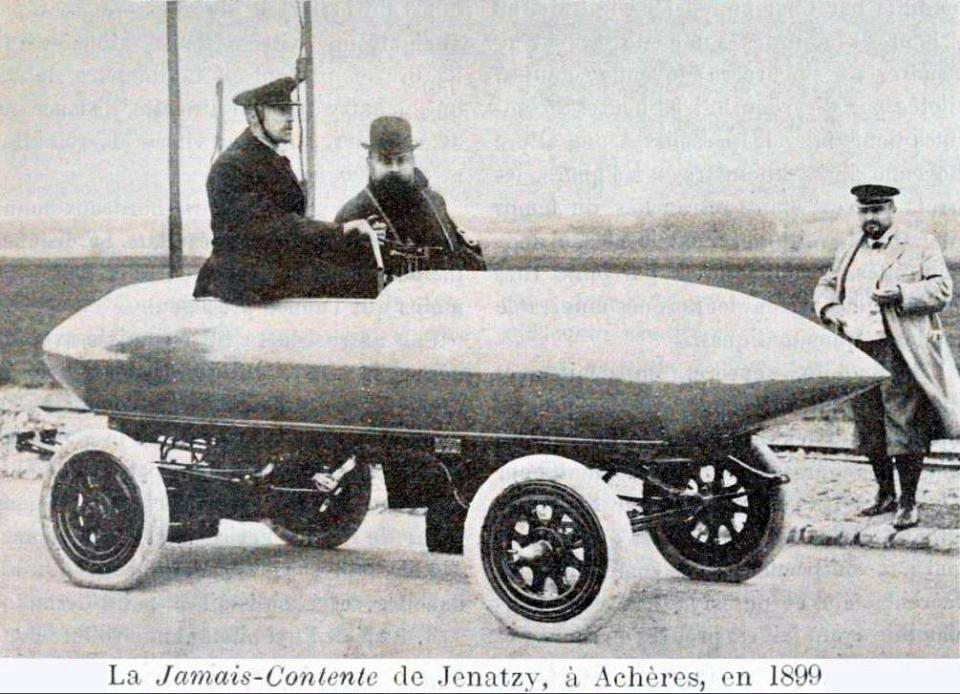 Photo en noir et blanc de la voiture Jamais contente, qui est en forme d'obus, avec un homme à bord et deux hommes autour.