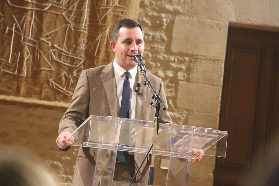 Un homme en costume beige parle à un pupitre. 