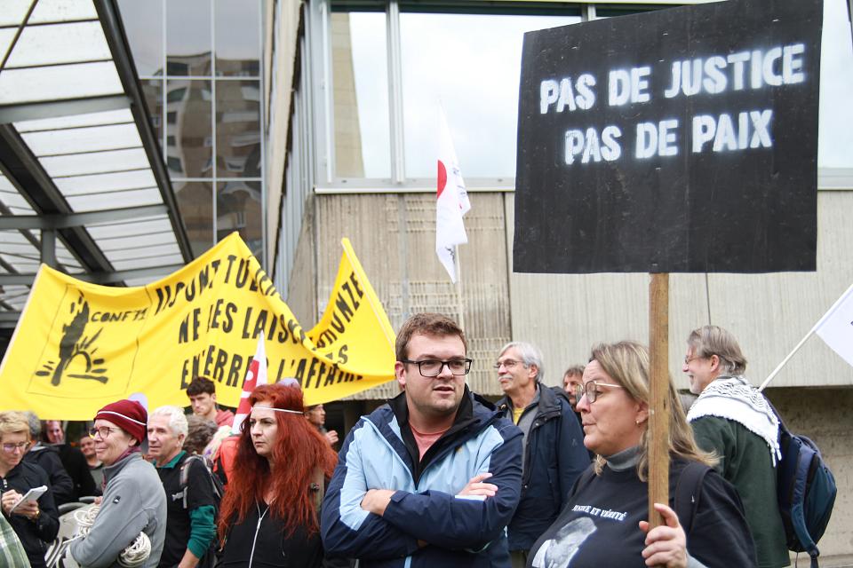 Une femme blonde avec des lunettes vêtue d'un t-shirt "Justice et vérité" avec le visage de Jérôme Laronze, porte une pancarte "pas de justice, pas de paix".