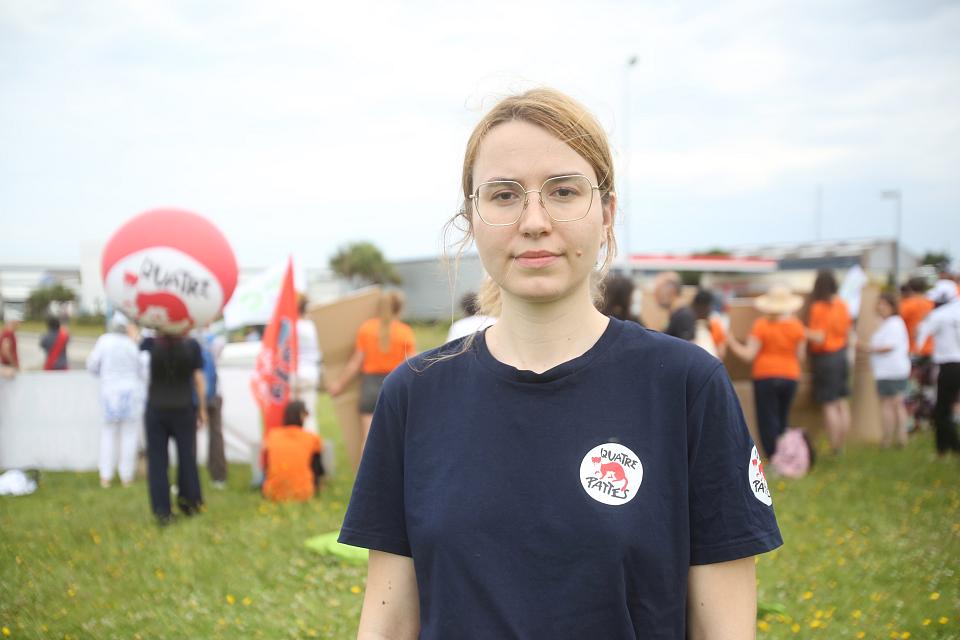 Une jeune femme regarde l'objectif, un groupe de personnes en t-shirt orange est visible dans le fonds dans un champs.