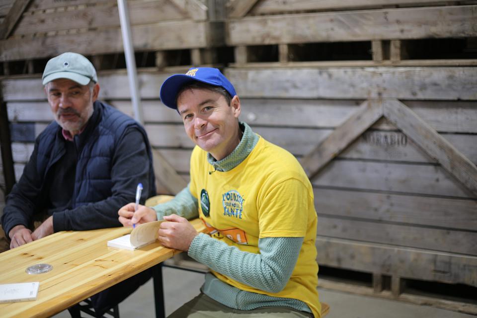 un homme avec un t-shirt jaune "l'odyssée de notre santé" et une casquette en train d'écrire, accoudé sur une table