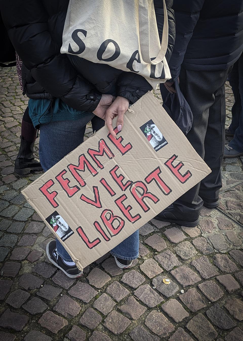 Une femme tient un carton sur lequel il est écrit "Femme, vie, liberté". On ne voit que ses mains et ses jambes. 