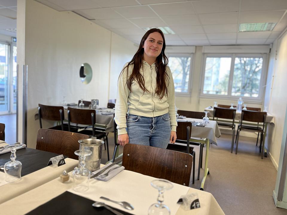 Une jeune femme, debout, entre des tables de restaurant dressées