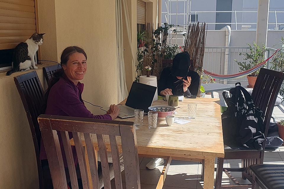 Une femme souriante assises à une table sur une terrasse, devant un ordinateur portable. Un chat se tient derrière elle.