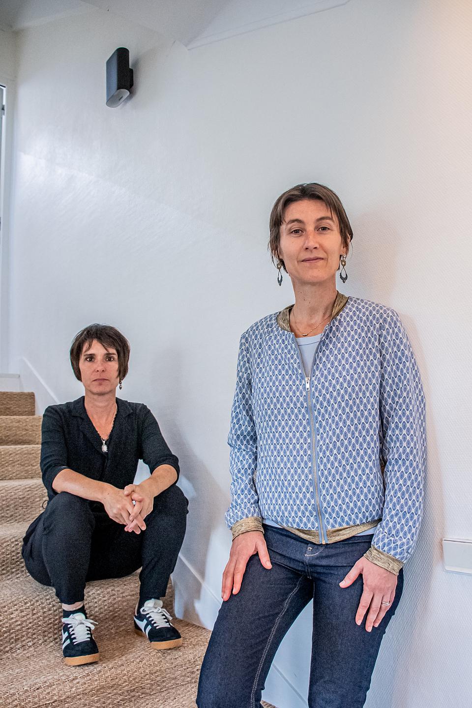 deux femmes, l'une vêtue de noir assise sur des escaliers, l'autre debout vêtue d'une veste bleue et dorée