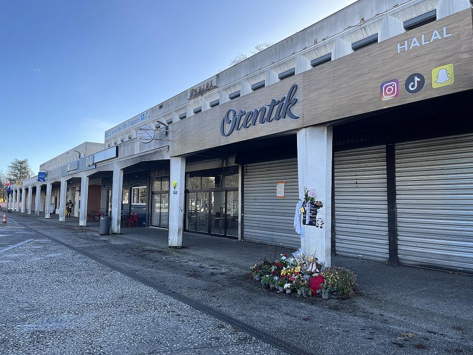 Une devanture de magasin dont le rideau est baissé. Au pied d'un pilier, on voit des fleurs.