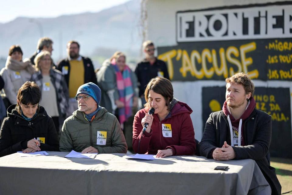 Quatre personnes sont assises à une table, une femme parle au micro. On voit des personnes debout derrière. 