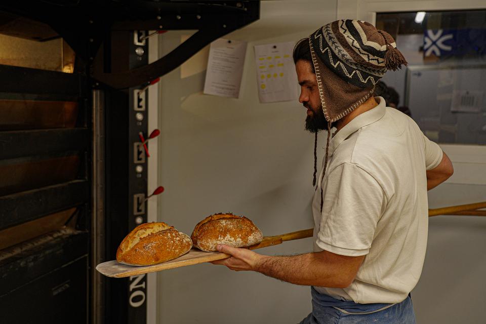 Un homme coiffé d'un bonnet en laine sort deux miches de pain d'un grand four. 