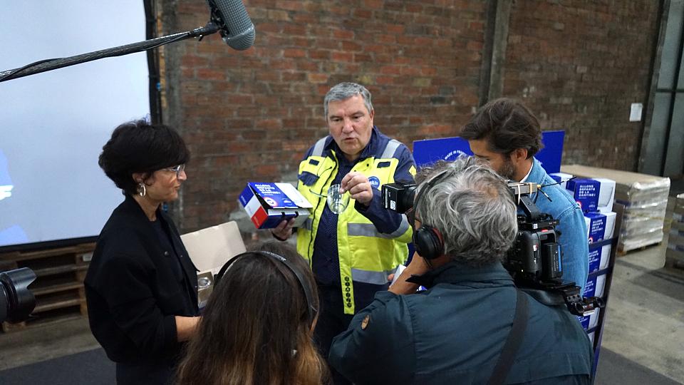 Un homme en chasuble jaune présente des modèles de verre à des journalistes qui tiennent des caméras et micro 