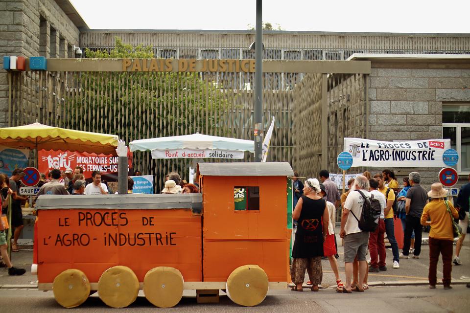Un rassemblement dans une rue : une trentaine de personnes debout, vues de dos, quelques banderoles. Au premier plan une fausse locomotive en bois est garée sur la route. Dessus, il est écrit "Le procès de l'agro-industrie".