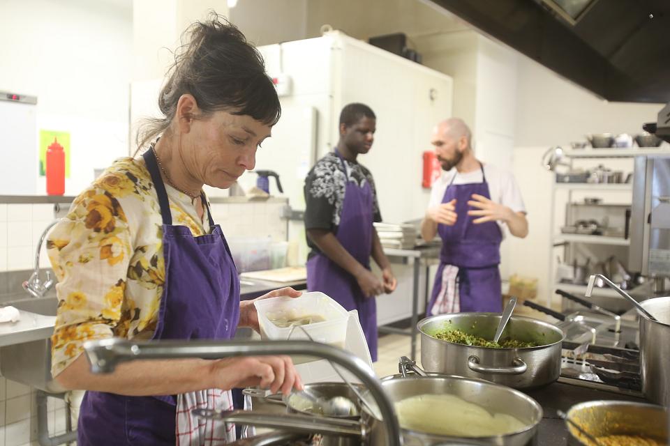 Une femme au dessus d'une casserole au premier plan, deux hommes parlent au second plan, dans une cuisine collective. Tous portent un tablier violet.