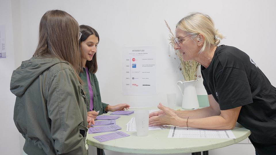 Deux jeunes femmes échangent avec une personne autour d'un table huate sur laquelle sont étalés des documents. 