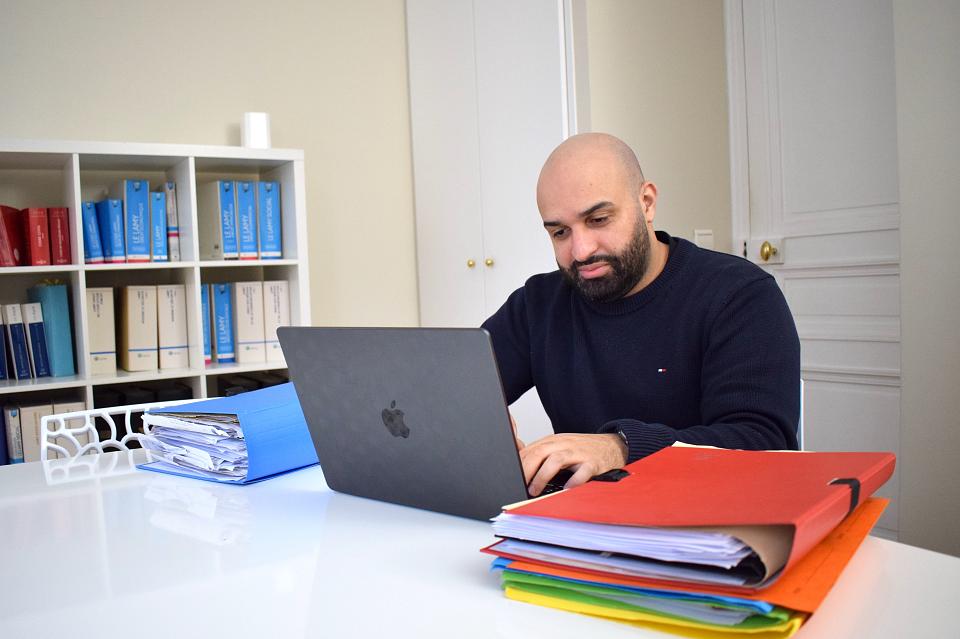 Un homme assis à un bureau devant un ordinateur portable, des dossiers sont posés sur le bureau.
