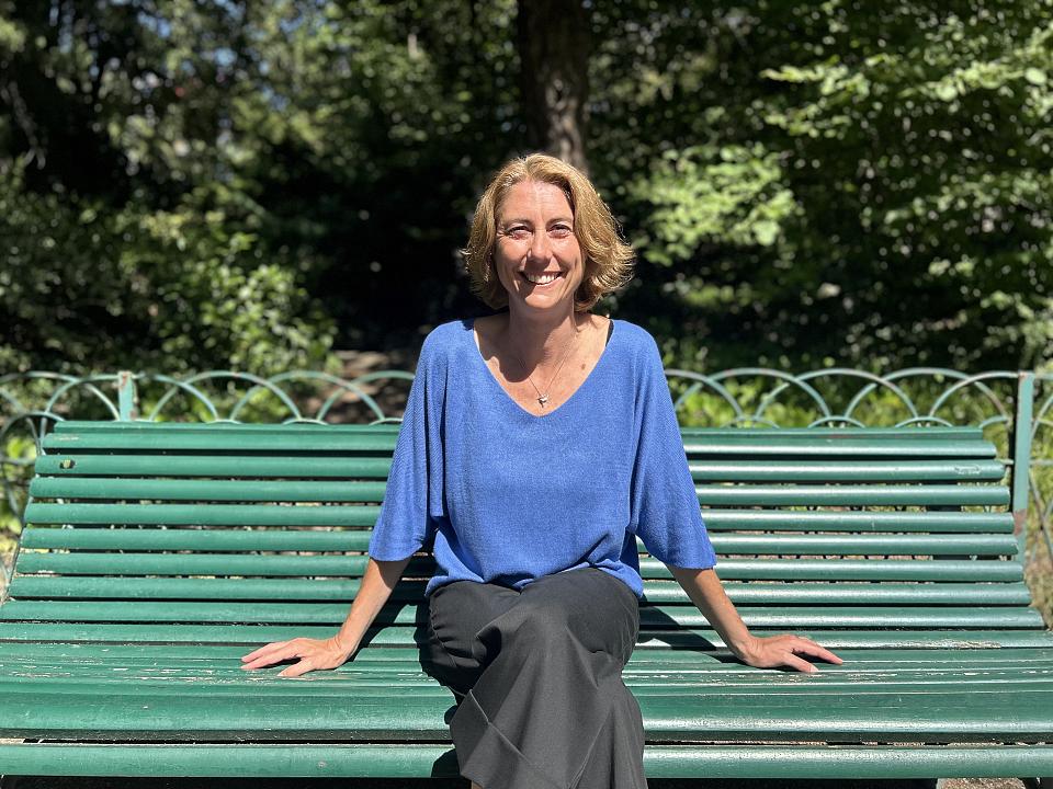 Une femme assise sur un banc dans un jardin public, elle regarde l'objectif en souriant