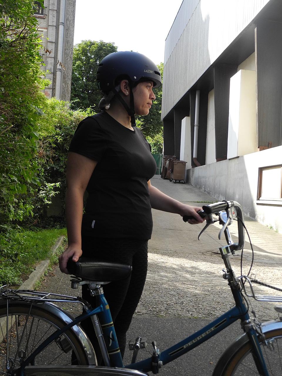 Une femme habillée en noir et de profil, porte un casque et tient un vélo bleu à côté d'elle.