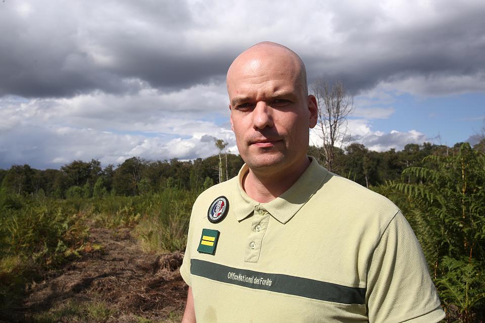 Un homme en t-shirt de l'ONF devant un paysage de forêt et un ciel gris. 