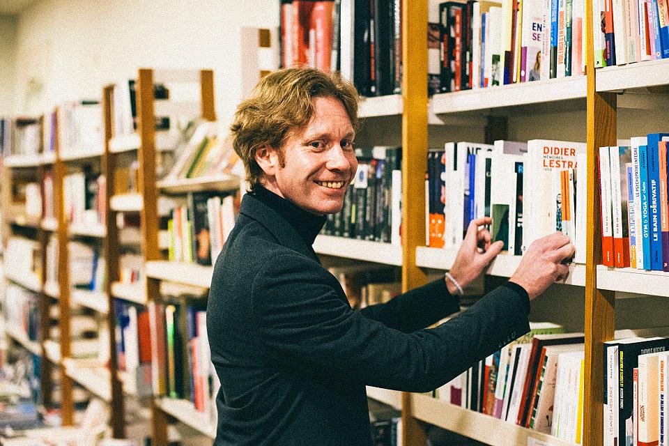 Un homme souriant devant des étagères de livres, il regarde l'objectif. 