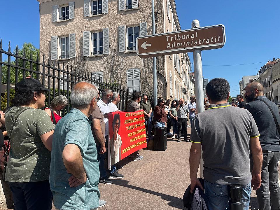 Un groupe de personnes avec une banderole rouge, à côté d'un panneau avec écrit "tribunal administratif". Une femme parle au micro.
