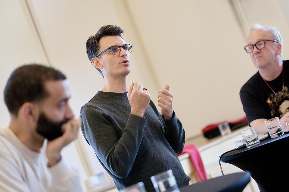Un homme parle dans ce qui semble être une conférence, entouré de deux autres qui écoutent