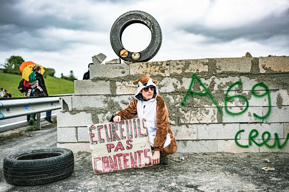 Une personne dans un costume d'écureuil est agenouillé devant un mir où il ets taggué A69 en vert et tient une pancarte sur laquelle il est écrit « écureuil pas contents ». Un pneu est posé à côté de lui. On voit des manifestant sur le bord d'une route dans le fond. 