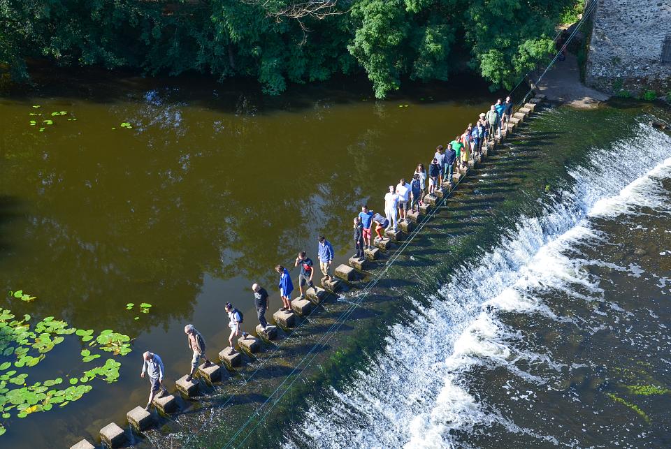 Une file de personnes vue du dessus traverse une rivière en marchand sur des pierres. 