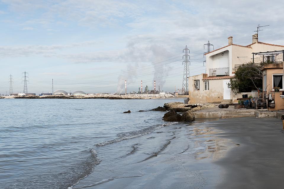 Vue sur la ZIP (zone industrialo-portuaire) de Fos-sur-mer depuis la plage jouxtant le port de plaisance. A droite des habitations, à gauche la mer, et sur la côte au fond, plusieurs usines fumantes et des lignes à haute tension. 