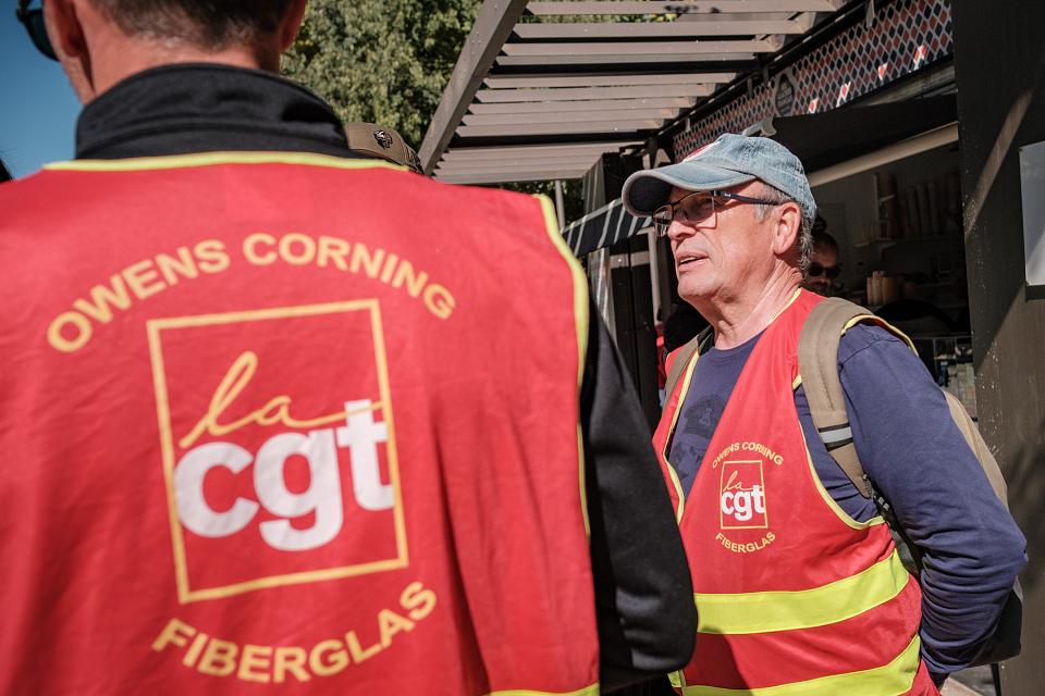 Un homme de profil porte un chasuble rouge de la CGT, un autre homme de dos porte aussi un chasuble rouge de la CGT sur lequel il est écrit « Owens Corning Fiberglas ».