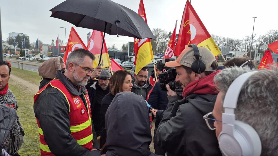Une femme parle avec des hommes en chasubles rouge et avec des drapeaux de la CGT, un homme filme. 