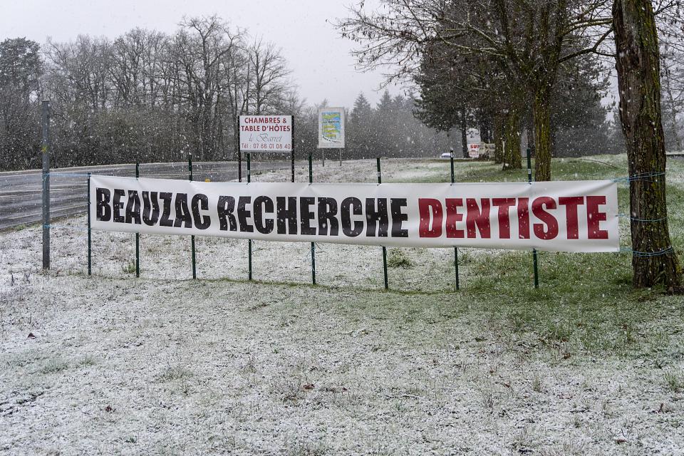 Une banderole sur laquelle il est écrit "Beauzac recherche dentiste" au bord d'une route dans un décor enneigé. 